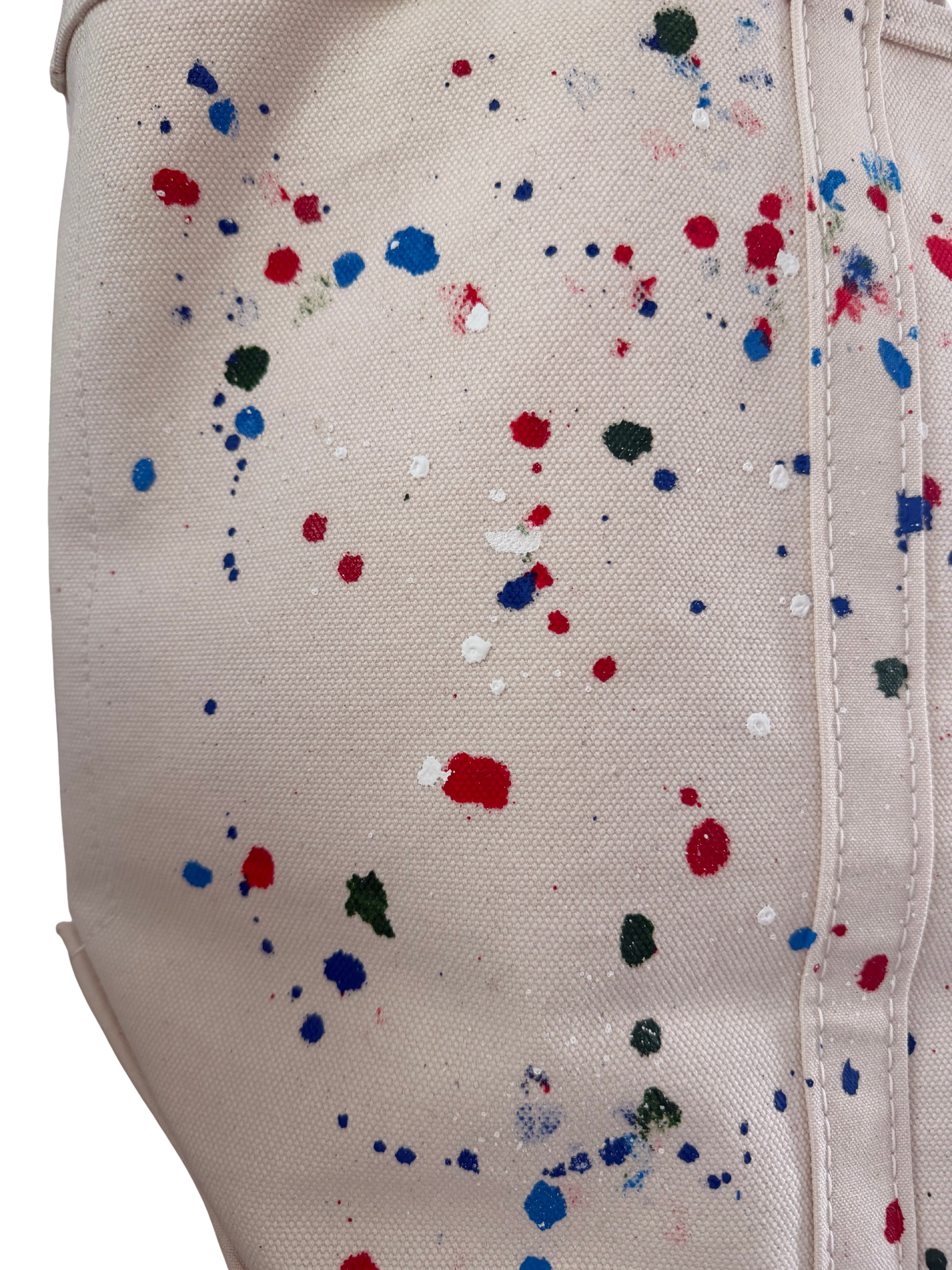 Close-up of a canvas tote bag in a warm off-white color, decorated with colorful paint splatters in red, blue, green, and white.