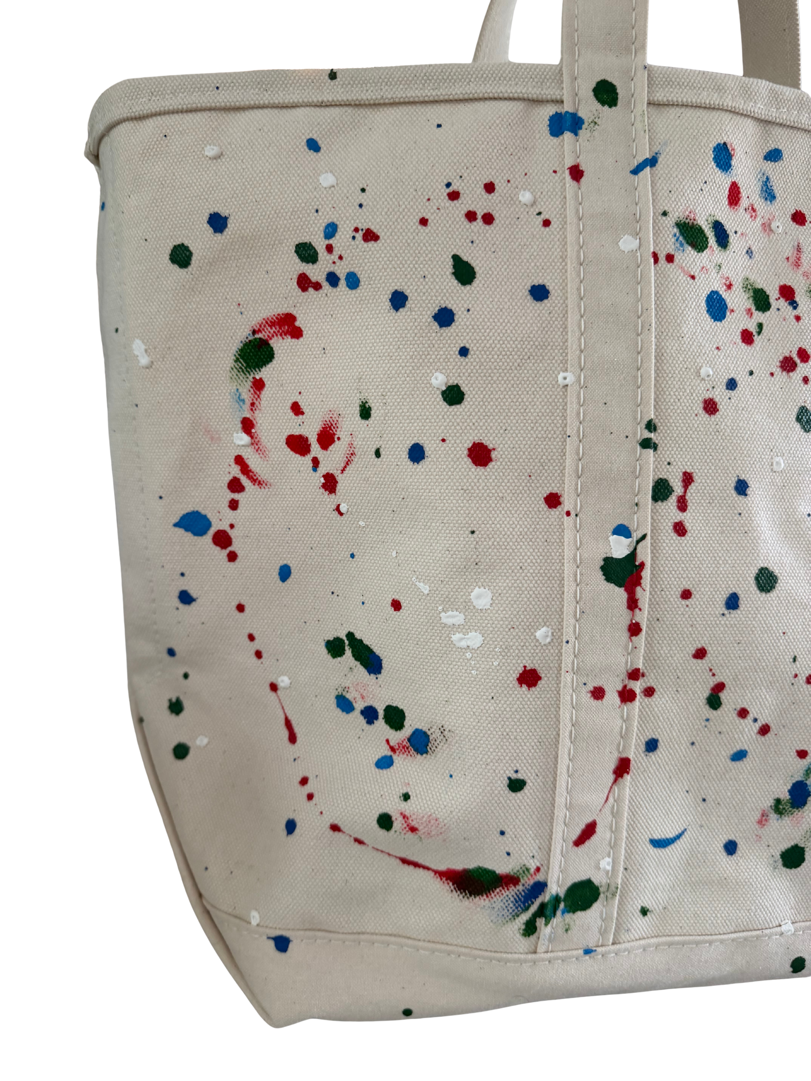 A cream canvas tote bag with colorful paint splatters in red, blue, green, and white, shown against a plain white background.