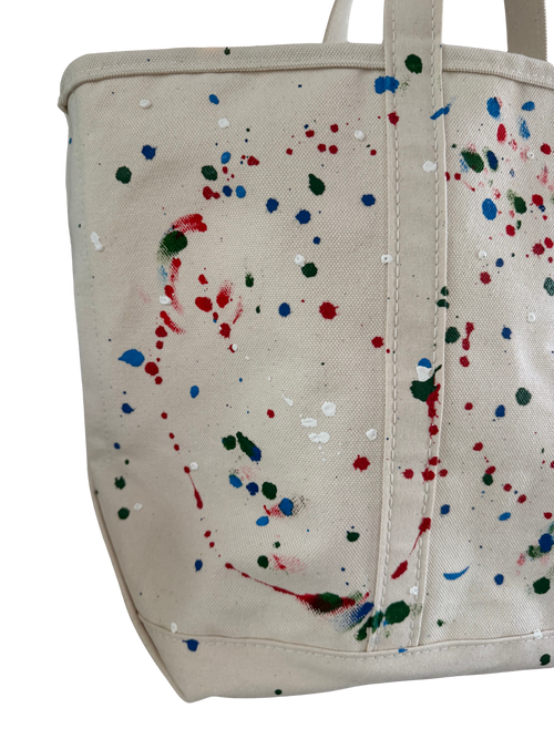 A cream canvas tote bag with colorful paint splatters in red, blue, green, and white, shown against a plain white background.