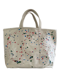 An off-white canvas tote bag with colorful paint splatters in red, blue, green, and white, featuring dual top handles, shown against a plain white background.