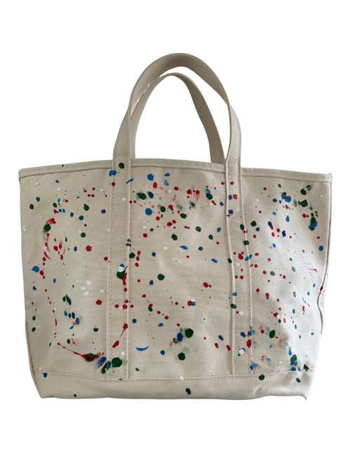 An off-white canvas tote bag with colorful paint splatters in red, blue, green, and white, featuring dual top handles, shown against a plain white background.